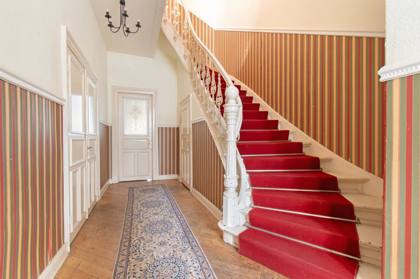 Grand foyer with a red-carpeted staircase, ornate white railing, patterned wallpaper, and a long decorative runner rug.