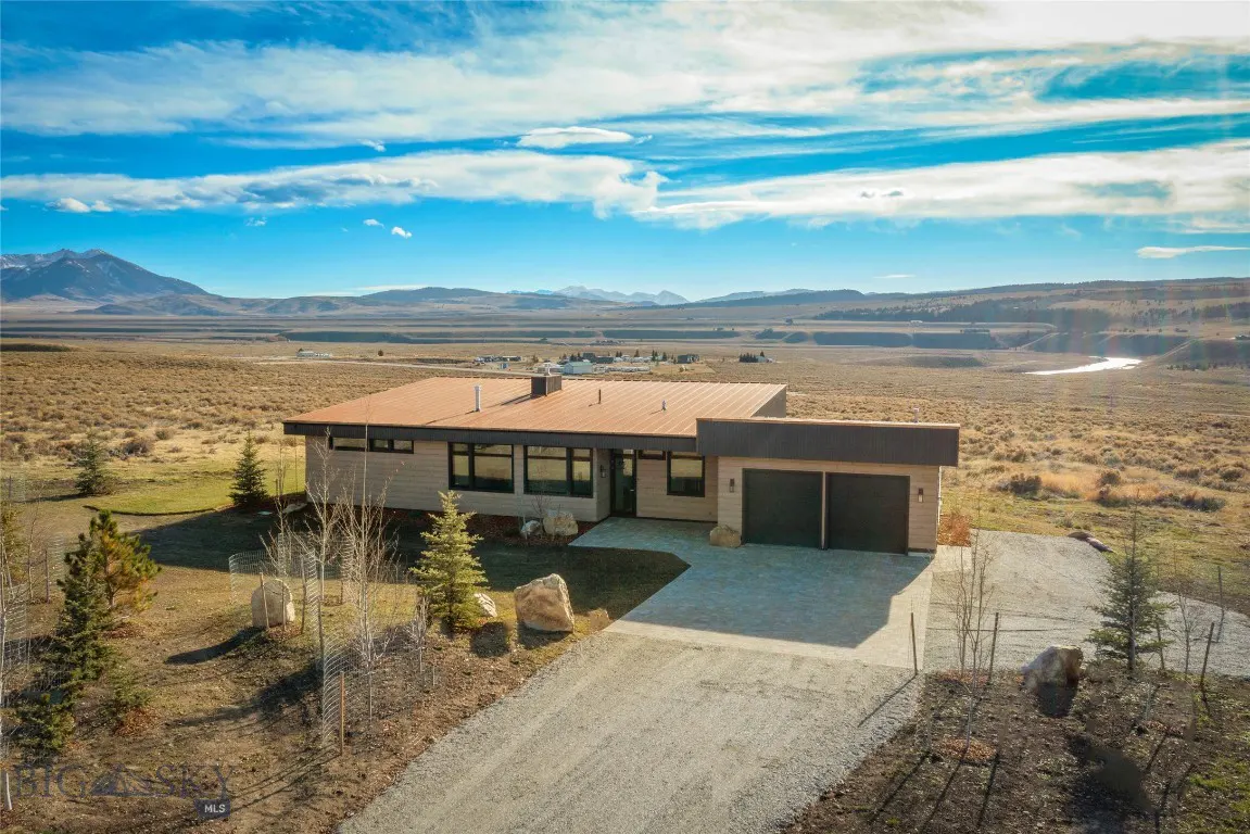 Modern beige house with a brown roof, a two-car garage, and a gravel driveway in a rural landscape with mountains in the background.