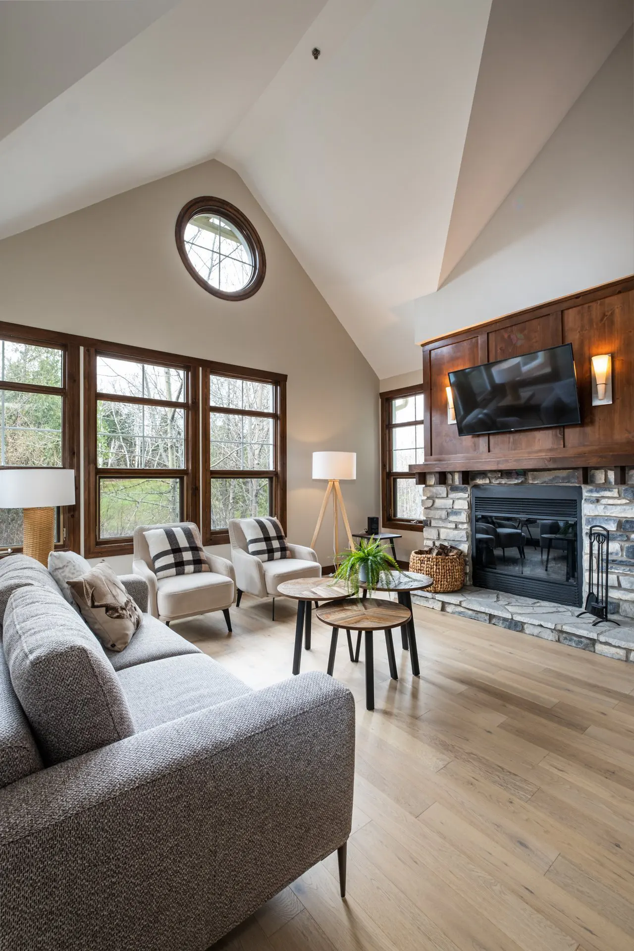 Living room with vaulted ceiling, wood floors, stone fireplace, and large windows. Gray sofa and two chairs face the fireplace.
