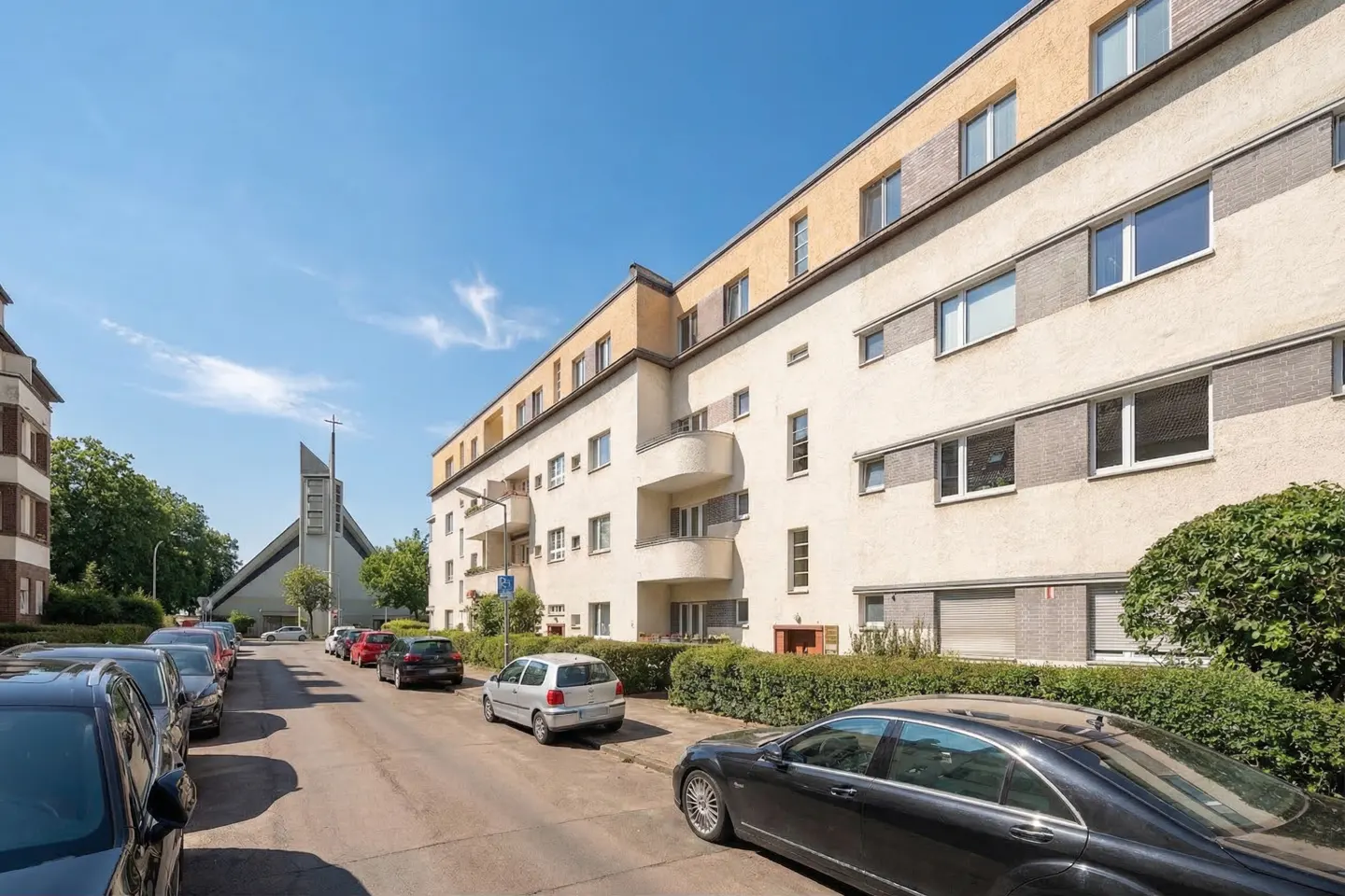 Street view of a cream apartment building with balconies, parked cars, and a church in the background.