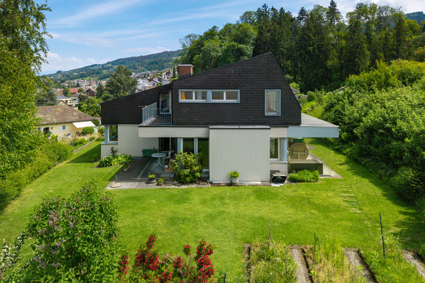 A modern two-story house with a dark roof and light siding, surrounded by a green lawn and trees.