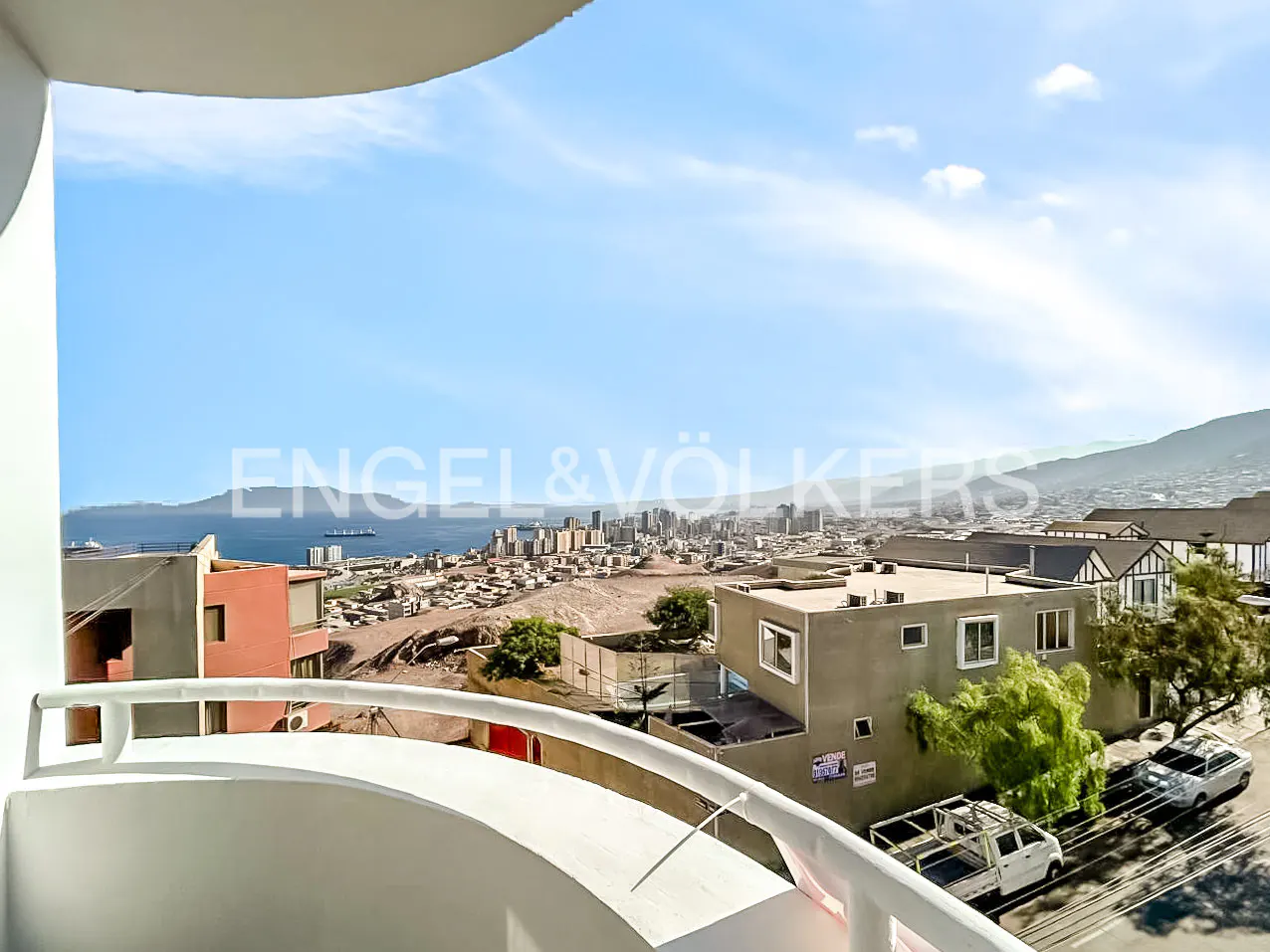 View from a white balcony overlooking a city, ocean, and mountains under a blue sky. Cars and buildings are visible.