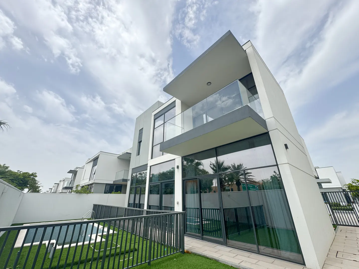 Modern white two-story house with a balcony and large windows, a pool, and a black metal fence.