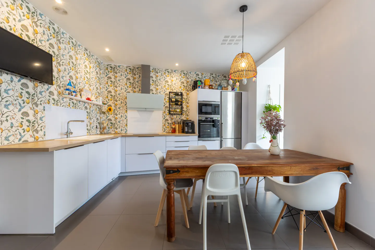 Bright kitchen with floral wallpaper, white cabinets, and a wooden table surrounded by white chairs. A woven pendant light hangs above the table.