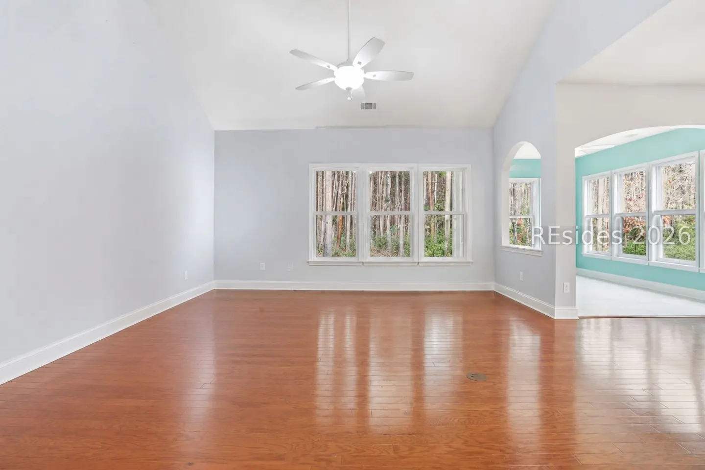 Empty living room with hardwood floors, light blue walls, and a white ceiling fan. Large windows offer a view of trees.