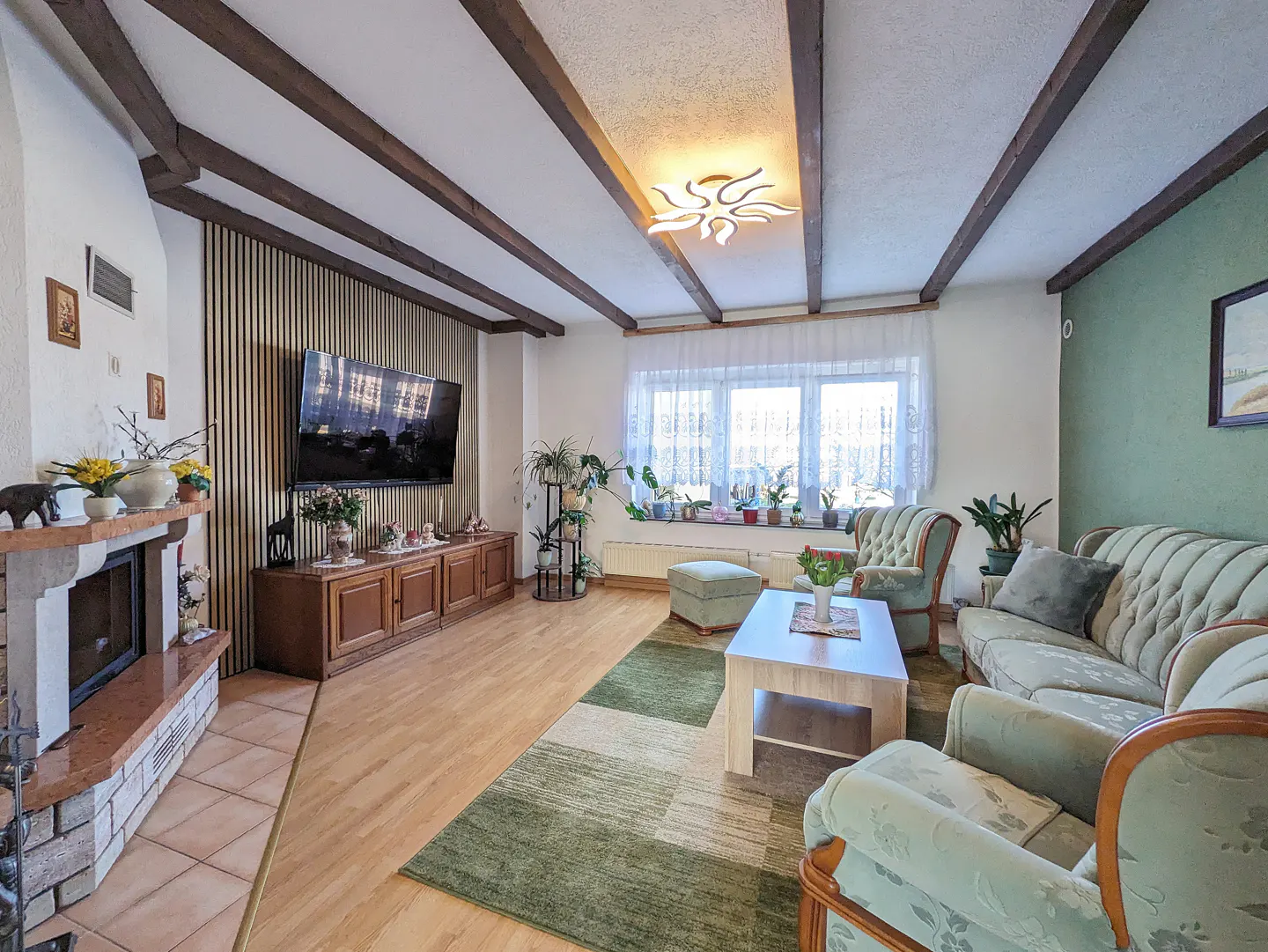 Living room with wood floors, exposed beams, and a stone fireplace. A green sofa and chairs face a coffee table and TV.