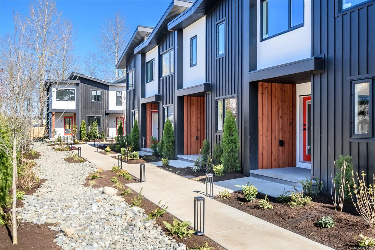 Row of modern townhouses with black siding, white accents, and red doors. A concrete walkway leads to each entrance, lined with landscaping and small lights.
