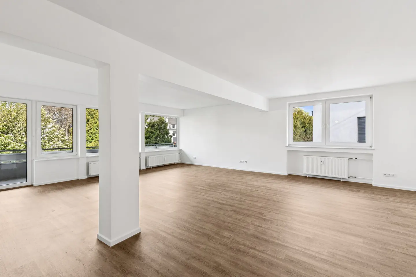 Bright, empty room with wood floors, white walls, and large windows showing green trees outside.