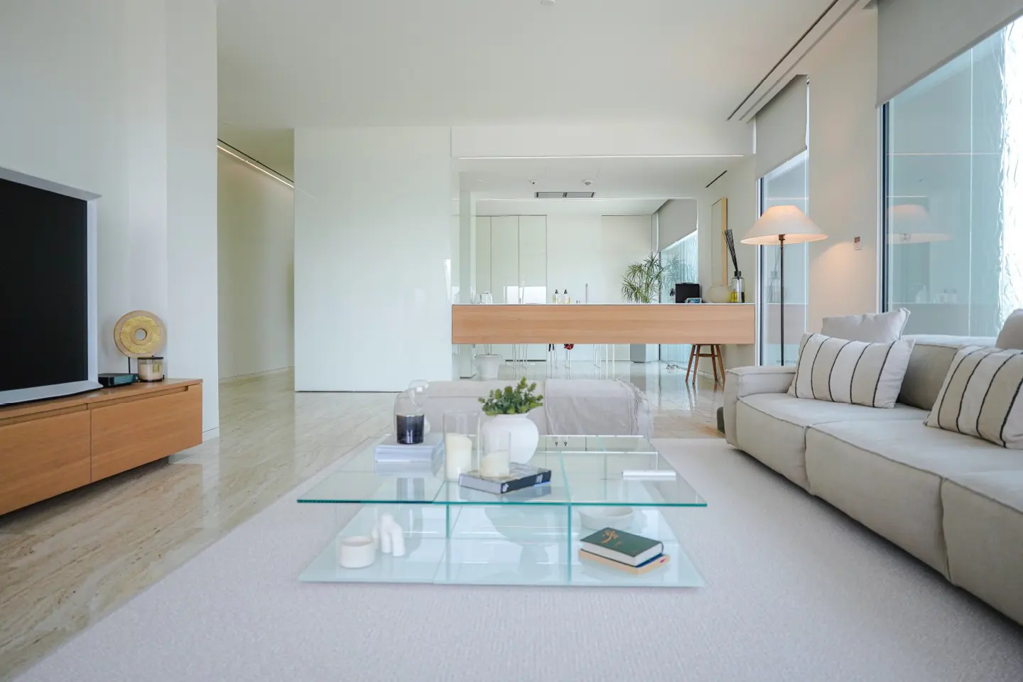 Bright, modern living room with a beige sofa, glass coffee table, and a TV on a wooden stand. White walls and large windows create an airy space.