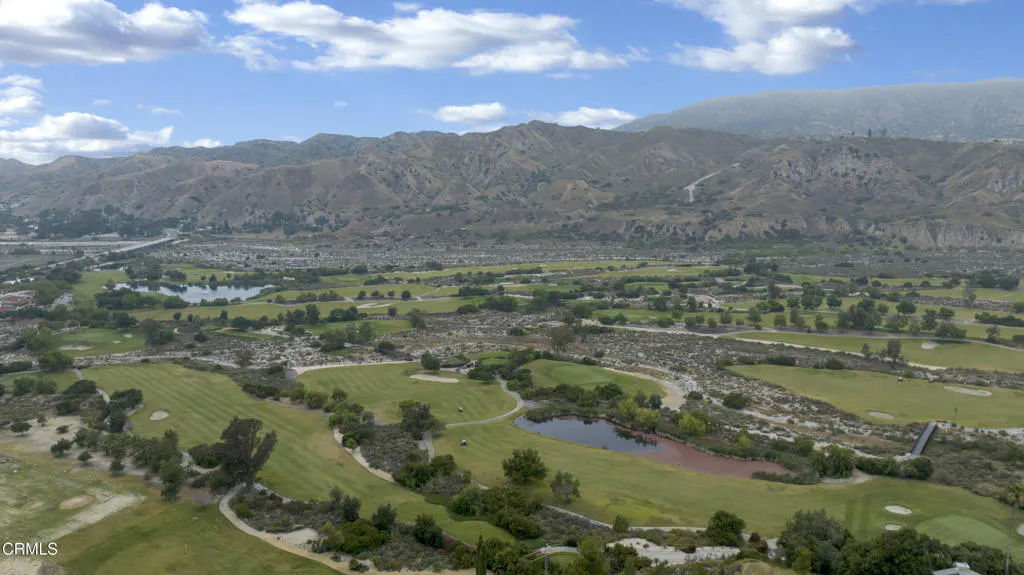 Aerial view of a green golf course with ponds, trees, and mountains in the background under a blue sky with white clouds.