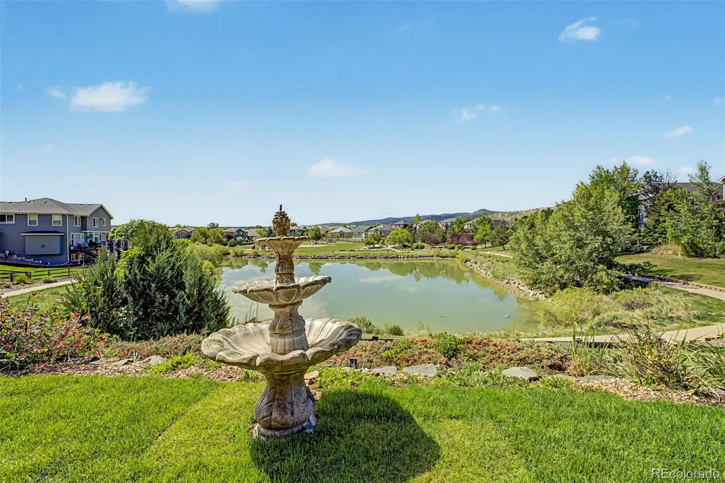 Stone fountain in a grassy yard overlooking a pond, suburban homes, and distant hills under a bright blue sky.