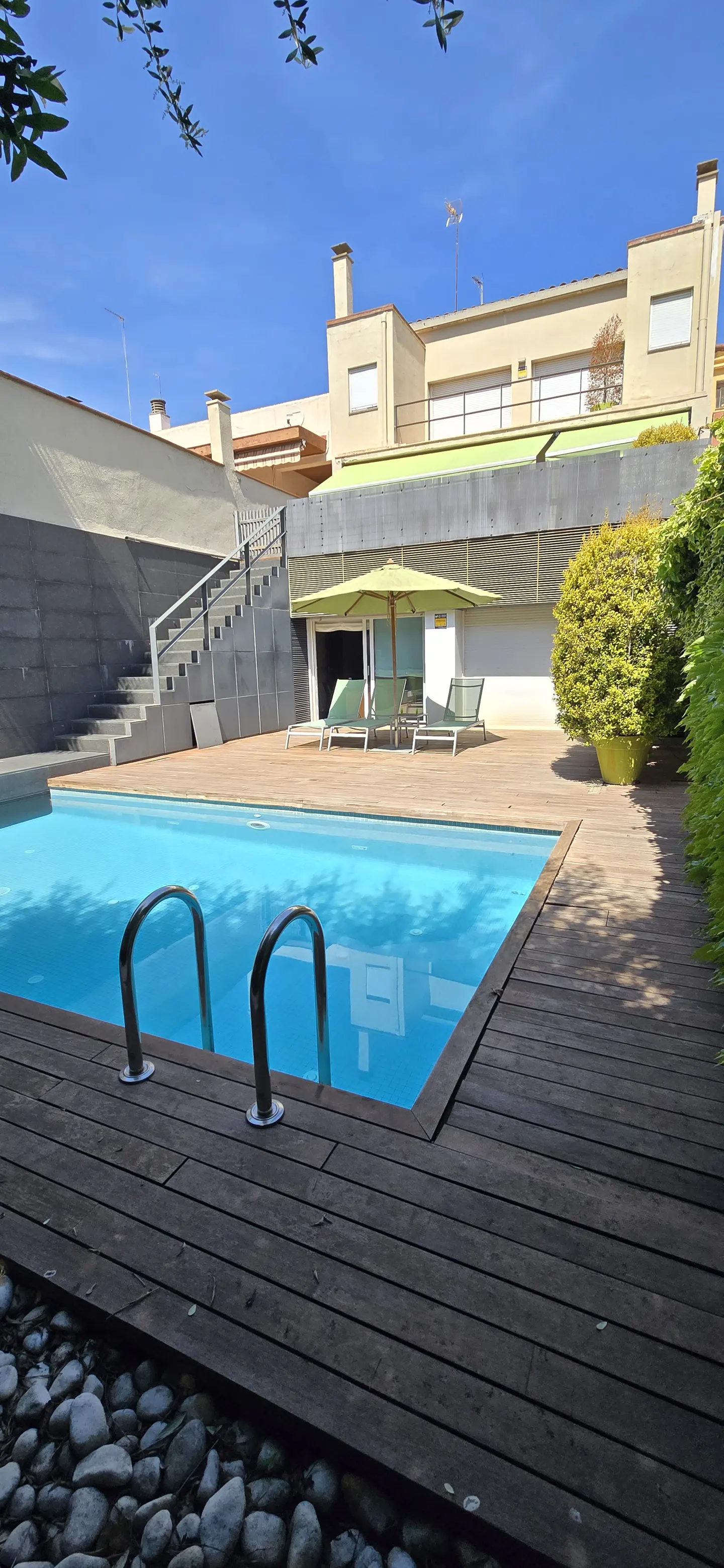 A backyard swimming pool with a wooden deck and ladder, lounge chairs, and a green umbrella.