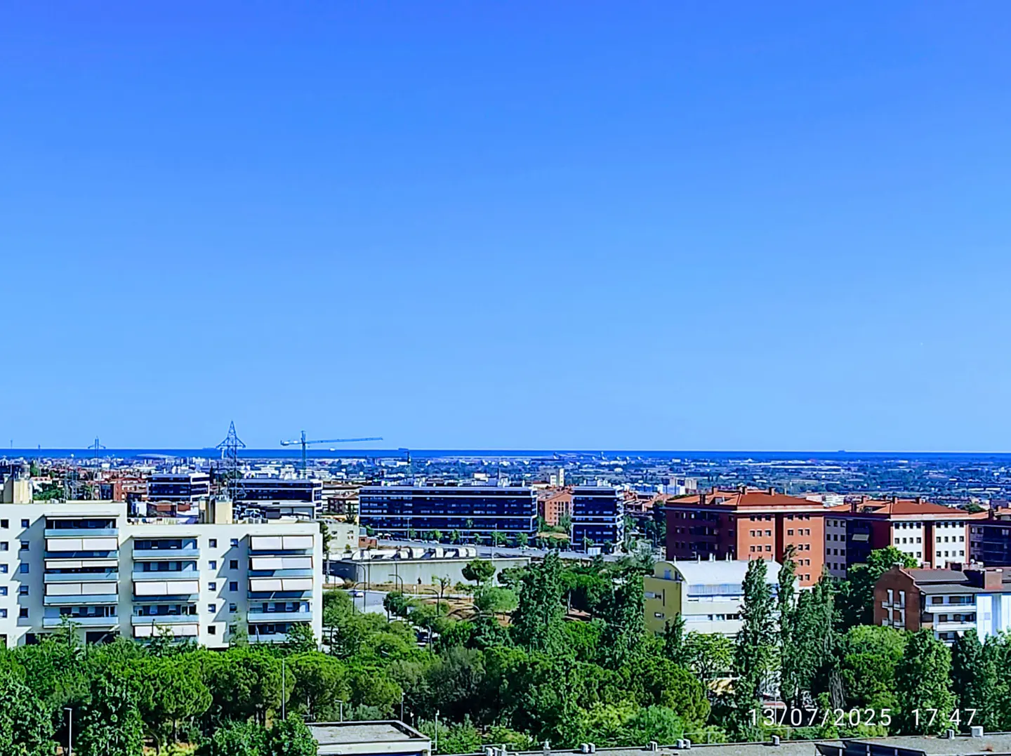 Cityscape view under a clear blue sky. Buildings, trees, and distant ocean visible.