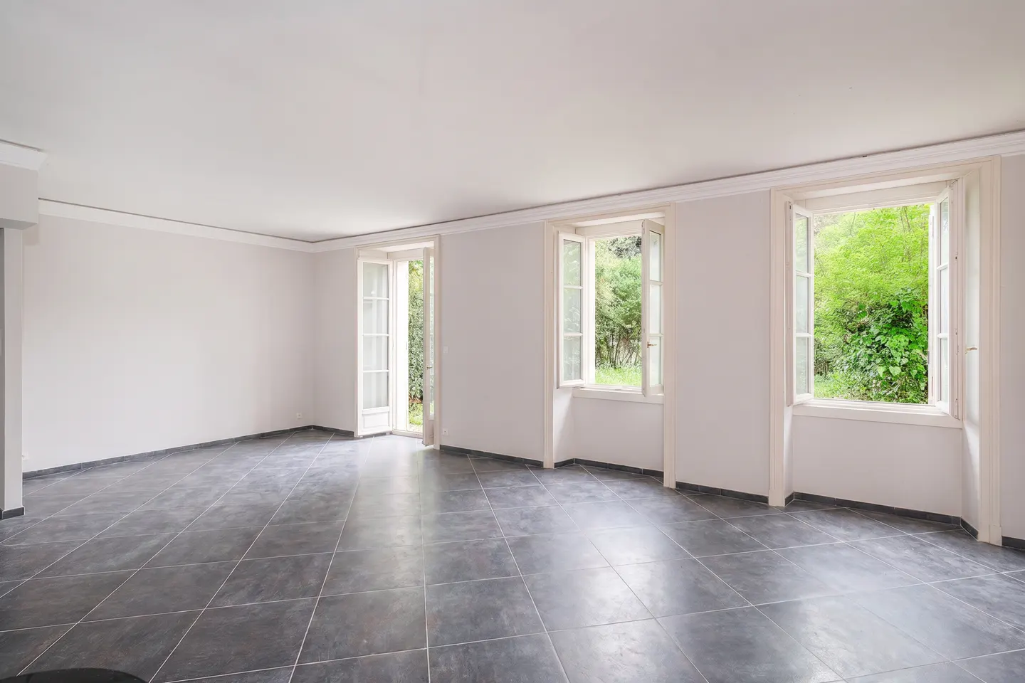 Bright, empty room with gray tile floor, white walls, and three open windows showing green foliage outside.
