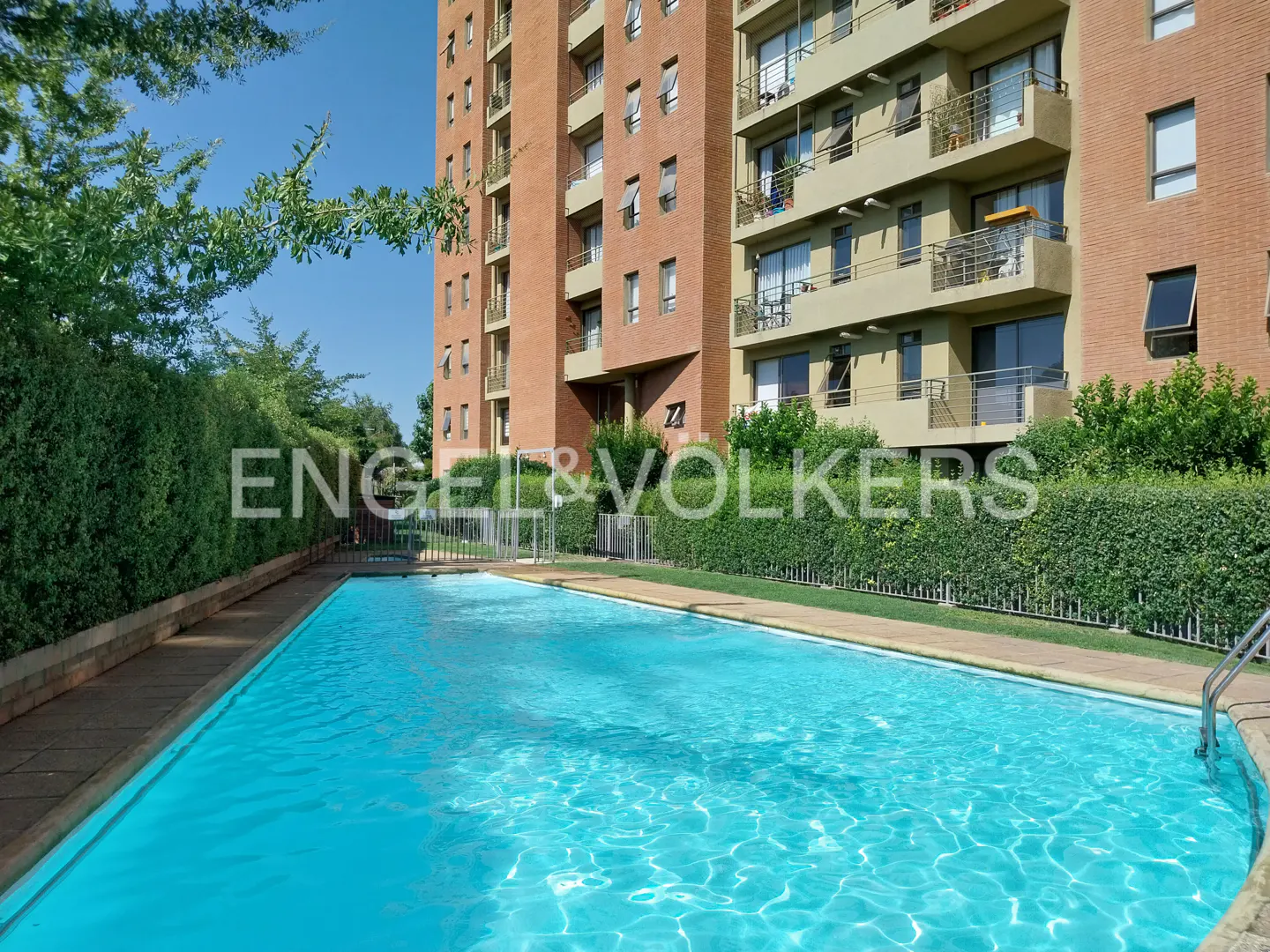 Outdoor pool with turquoise water in front of a brick apartment building with balconies and the Engel & Volkers logo.