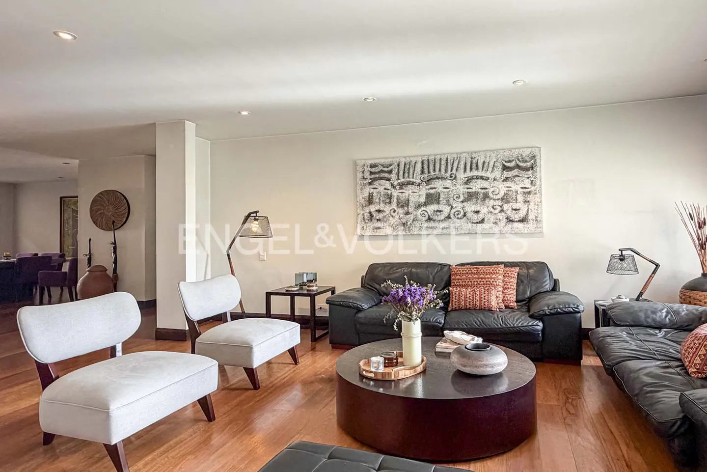 Modern living room with white chairs, black leather sofas, a round coffee table, and abstract wall art on hardwood floors.