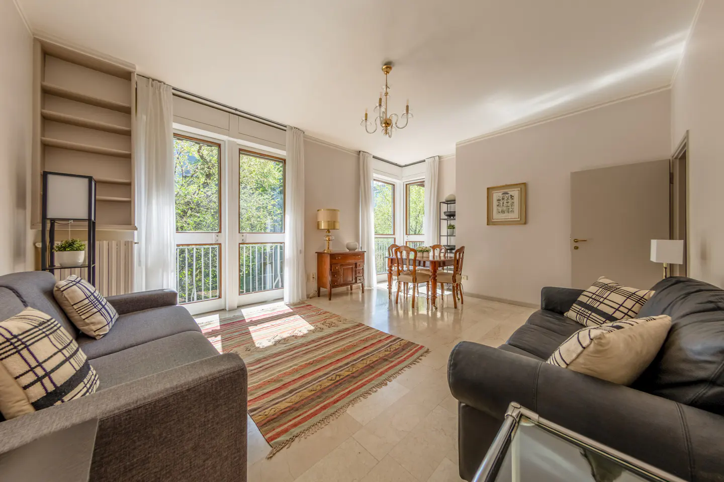 Bright living room with gray sofas, plaid pillows, and a striped rug. Dining table and balcony doors offer a view of lush greenery.