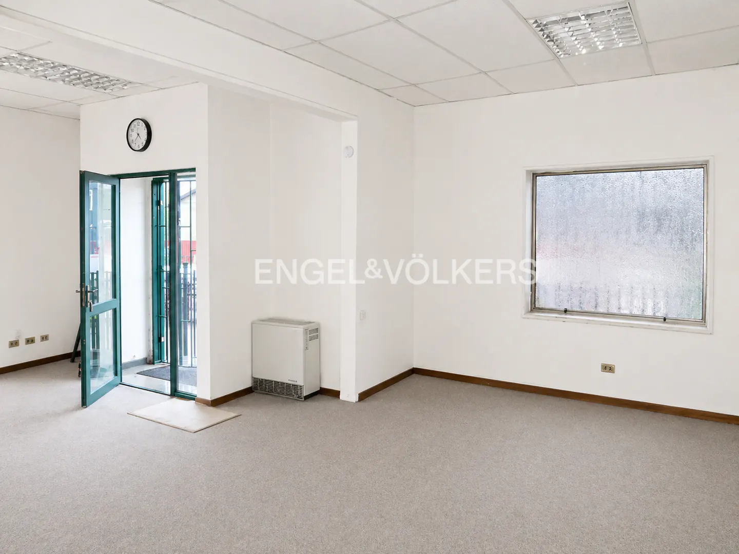 Bright, empty room with gray carpet, white walls, and a window. A green-framed glass door is open to the left, with a clock above.