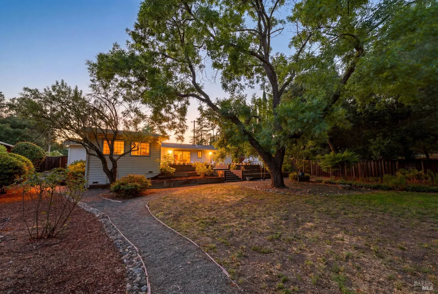 A gravel path leads to a house with warm interior lighting at dusk, surrounded by large trees and a landscaped yard.