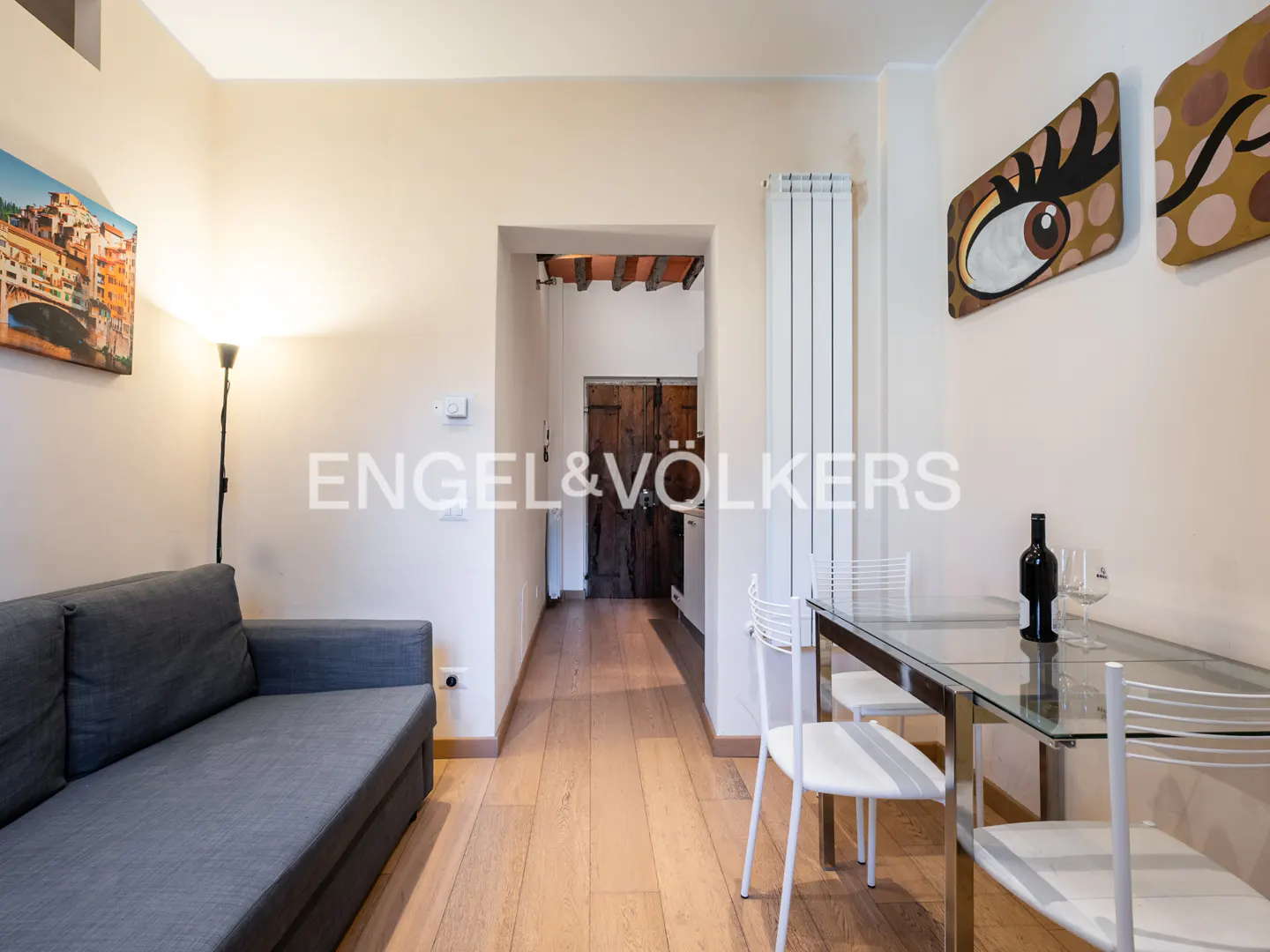 A living area with a grey sofa, a glass dining table with white chairs, and a doorway leading to a rustic wooden door.