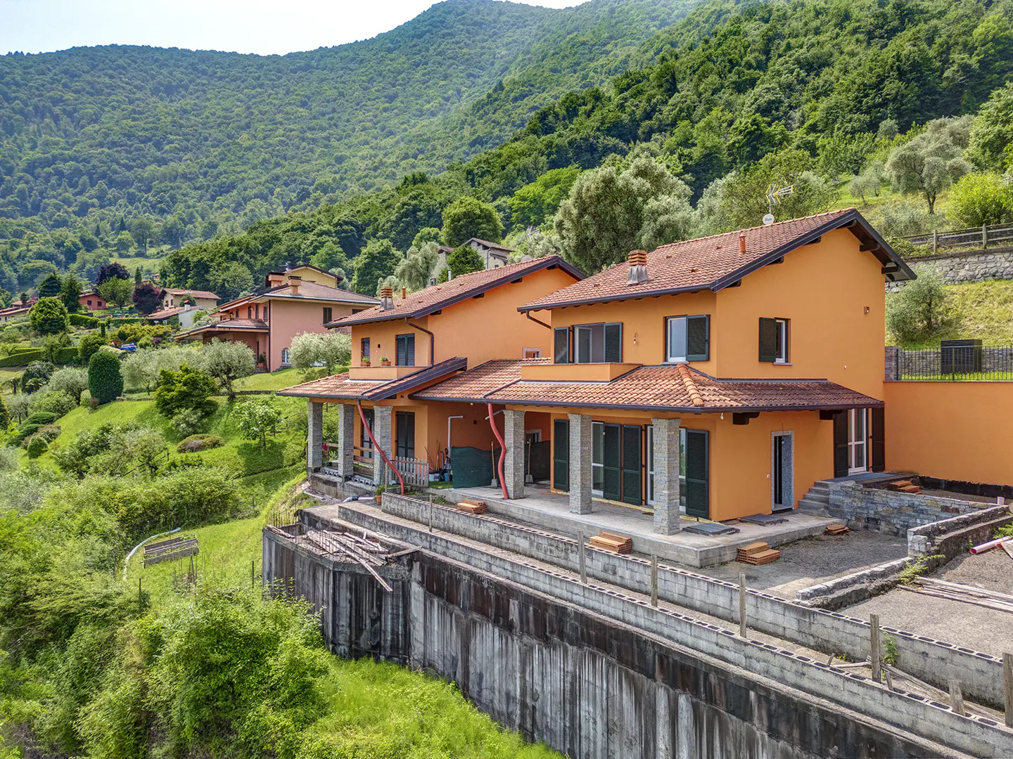 Orange two-story house with a red tile roof, set on a hillside with a lush green mountain backdrop.