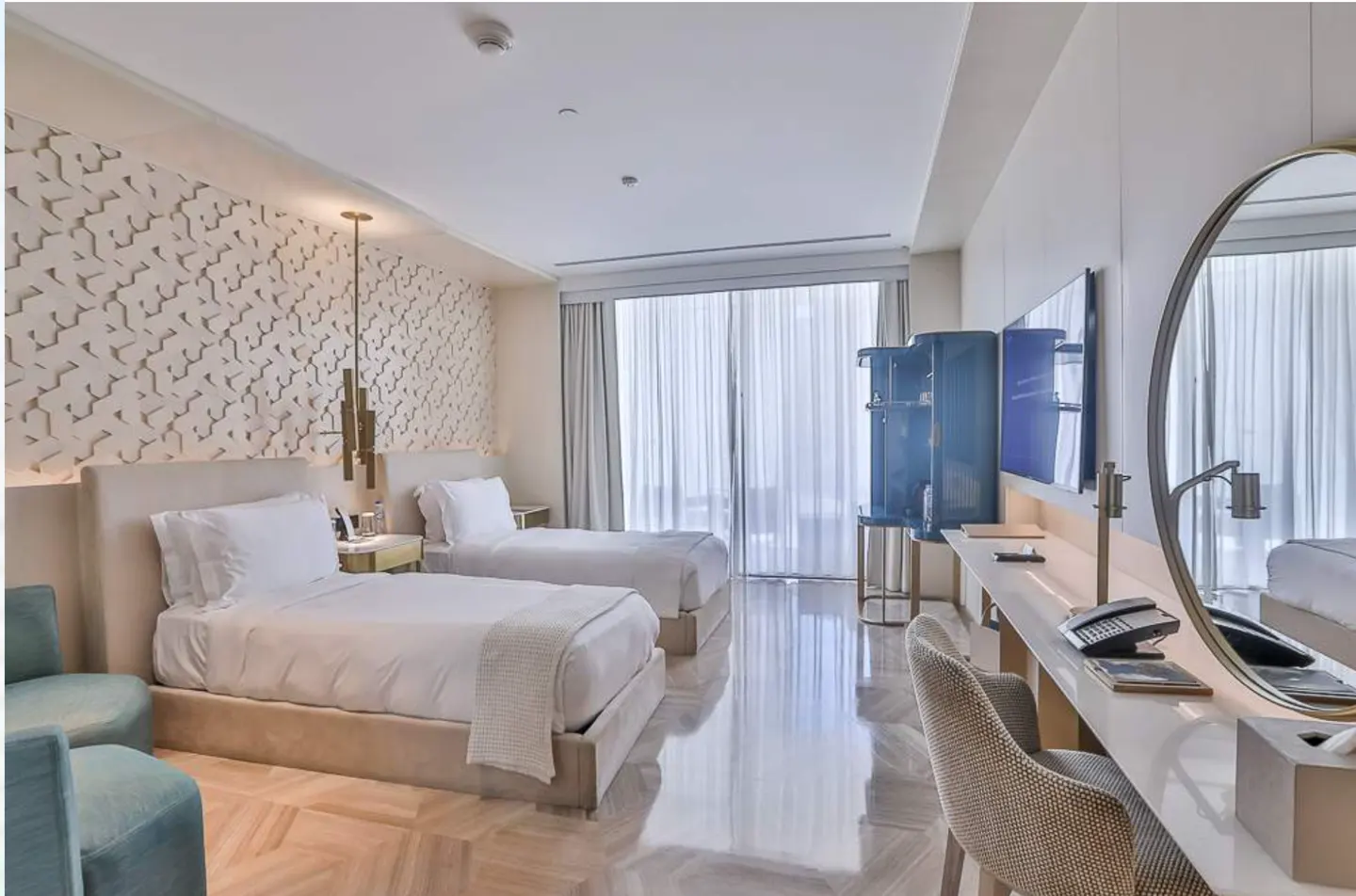 Bright hotel room with two twin beds, white linens, and textured wall. A blue cabinet and desk with a large mirror are on the right.