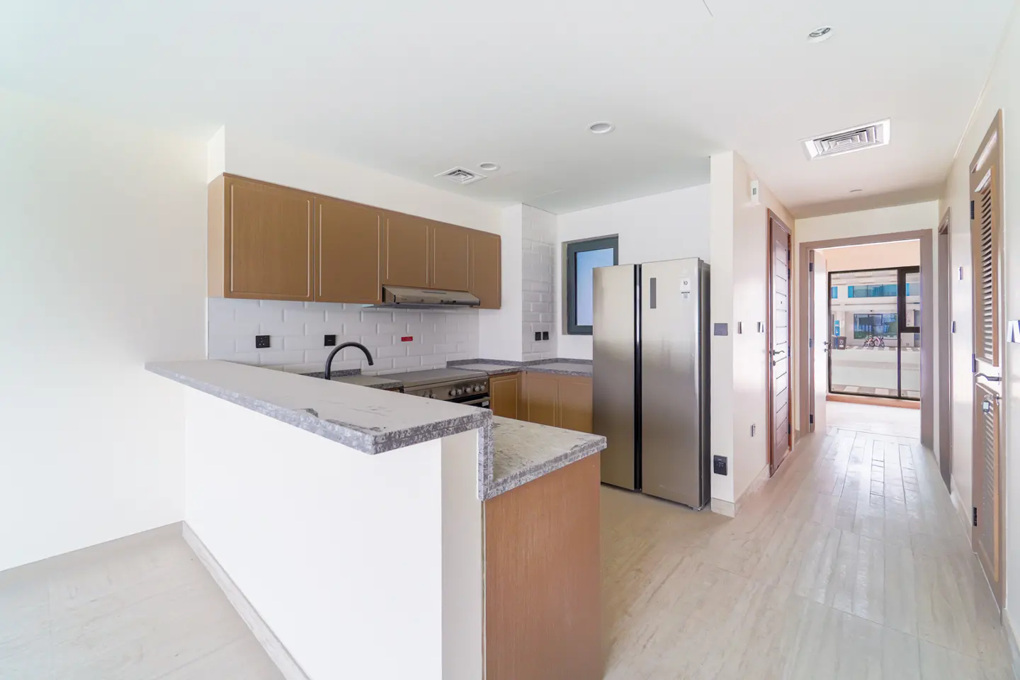 A bright, modern kitchen with brown cabinets, stainless steel appliances, and a breakfast bar. A hallway leads to a patio.
