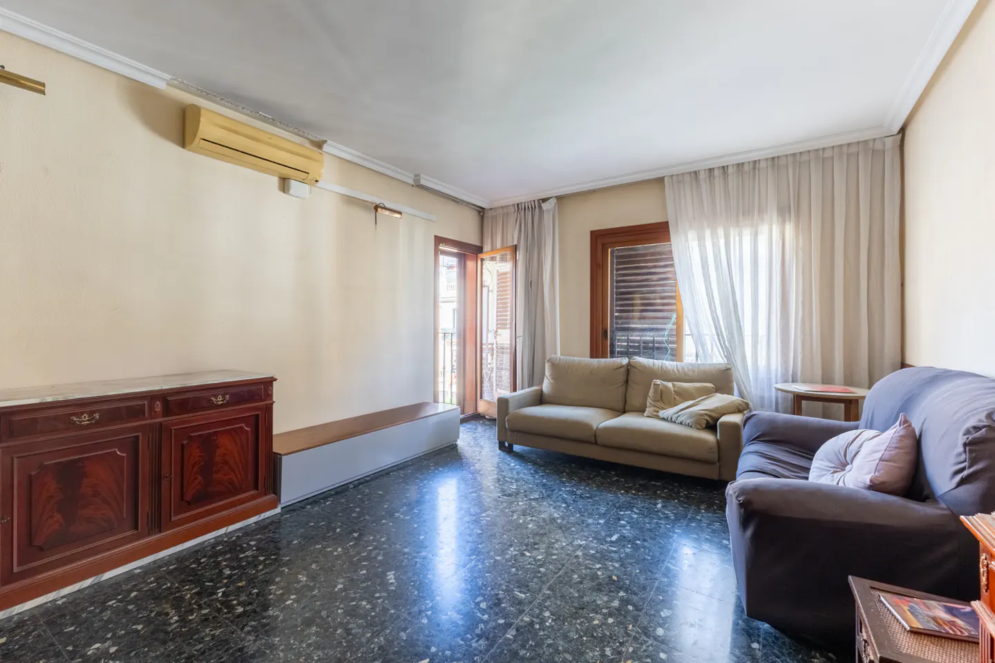 Living room with dark marble floors, a beige sofa, a dark armchair, a wooden cabinet, and sheer curtains over a balcony door.