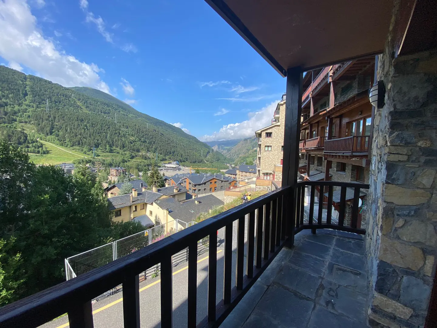 View from a stone balcony with a dark wood railing overlooking a town nestled in a green valley under a blue sky.