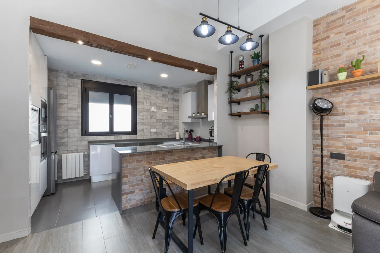 A modern kitchen with a wooden table and black chairs, brick walls, and industrial-style lighting. Shelves with plants add a touch of greenery.