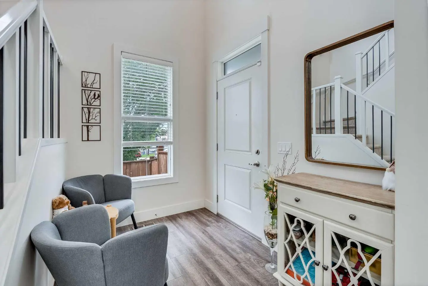 Bright entryway with gray chairs, white walls, and wood-look floors. A white door and window are visible, along with a cabinet and mirror.