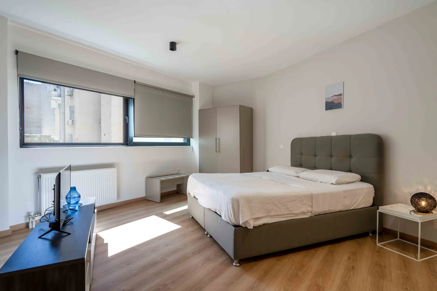 Bright bedroom with a gray bed, white linens, and wood floors. A large window with gray blinds overlooks a city view. A TV and wardrobe are also visible.