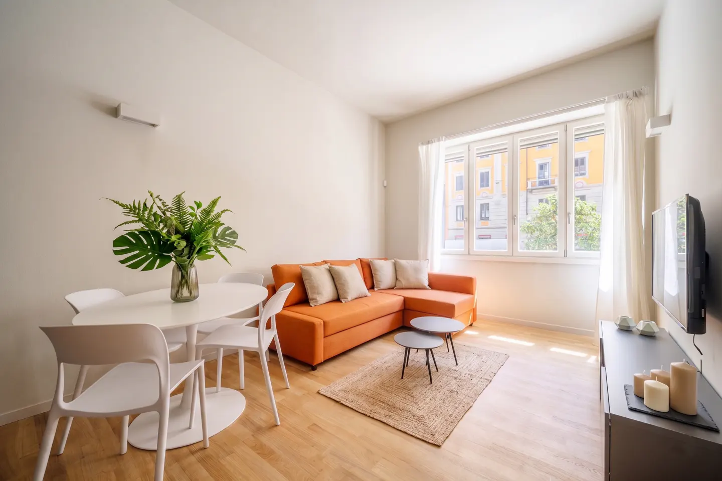 Bright living room with orange sofa, white table and chairs, and a large window with sheer curtains. A TV and console table are on the right.