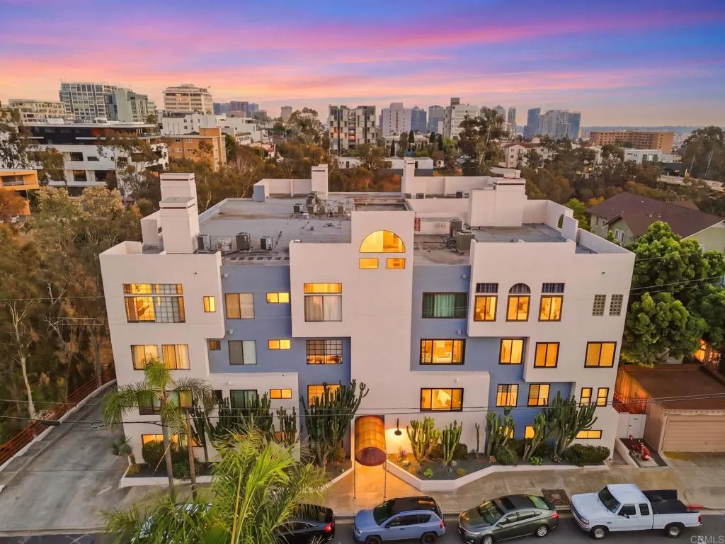 Exterior view of a modern, multi-story apartment building with blue and white facade, palm trees, and parked cars at dusk.