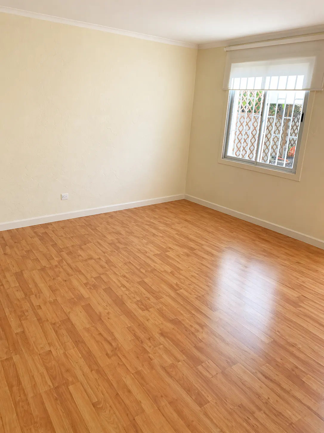 Empty room with light yellow walls and wood-look flooring. A window with white bars and a shade is on the right wall.
