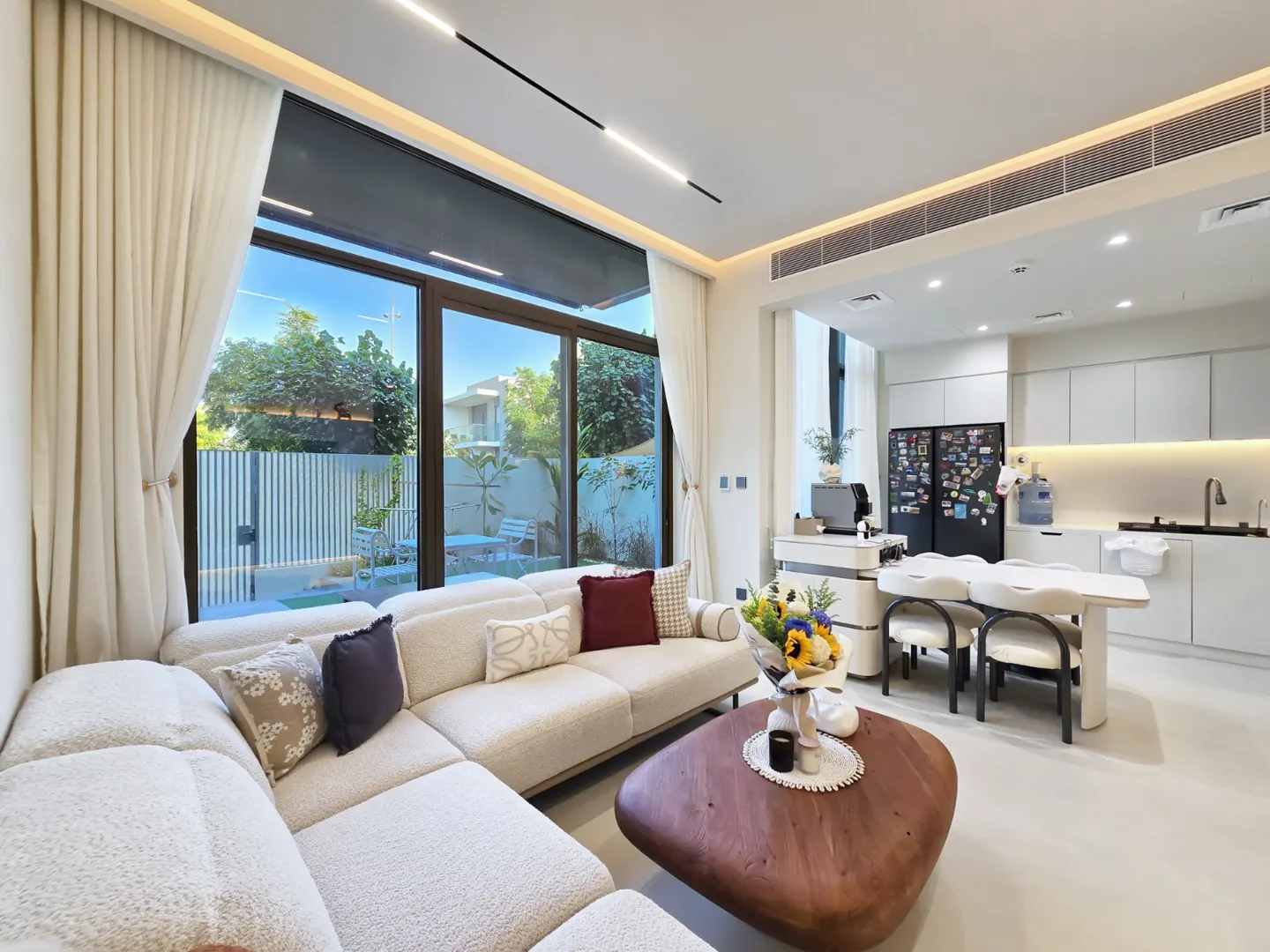 Bright, modern living room with a white sectional sofa, wood coffee table, and large windows overlooking a green backyard. White kitchen in the background.