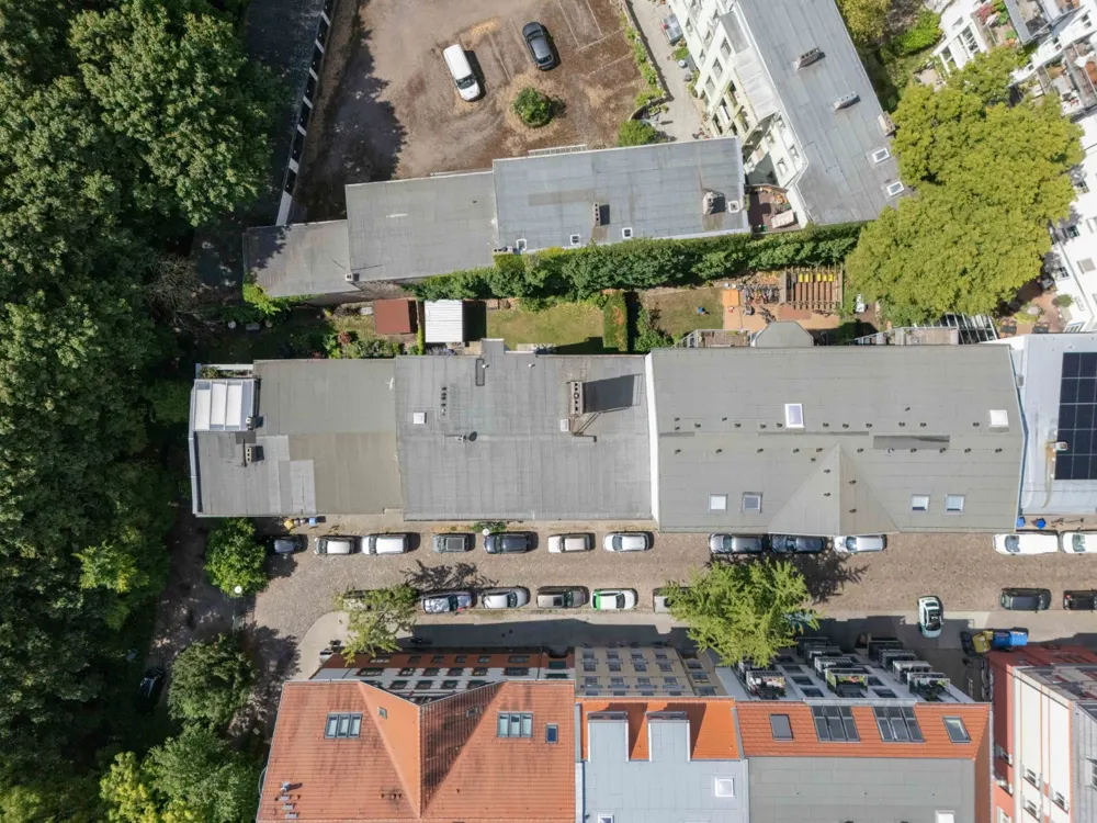 Aerial view of a city street with parked cars, buildings with gray and red roofs, and green trees.