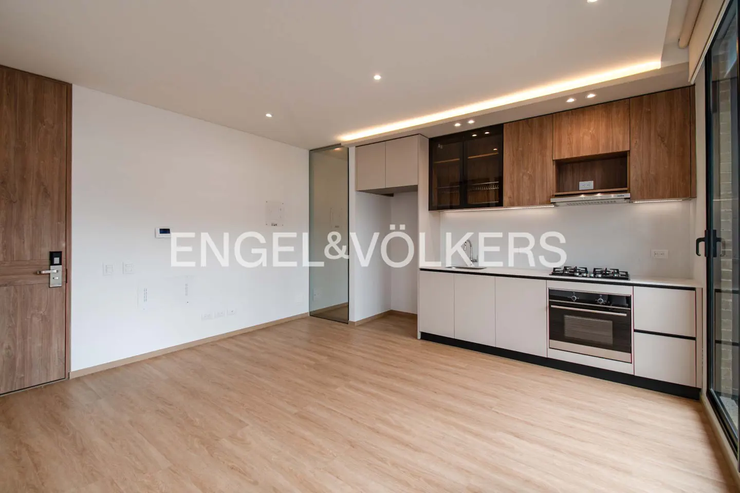 A bright, modern kitchen with wood floors, white walls, and two-tone cabinets. An oven and stovetop are visible. Engel & Völkers logo is overlaid.