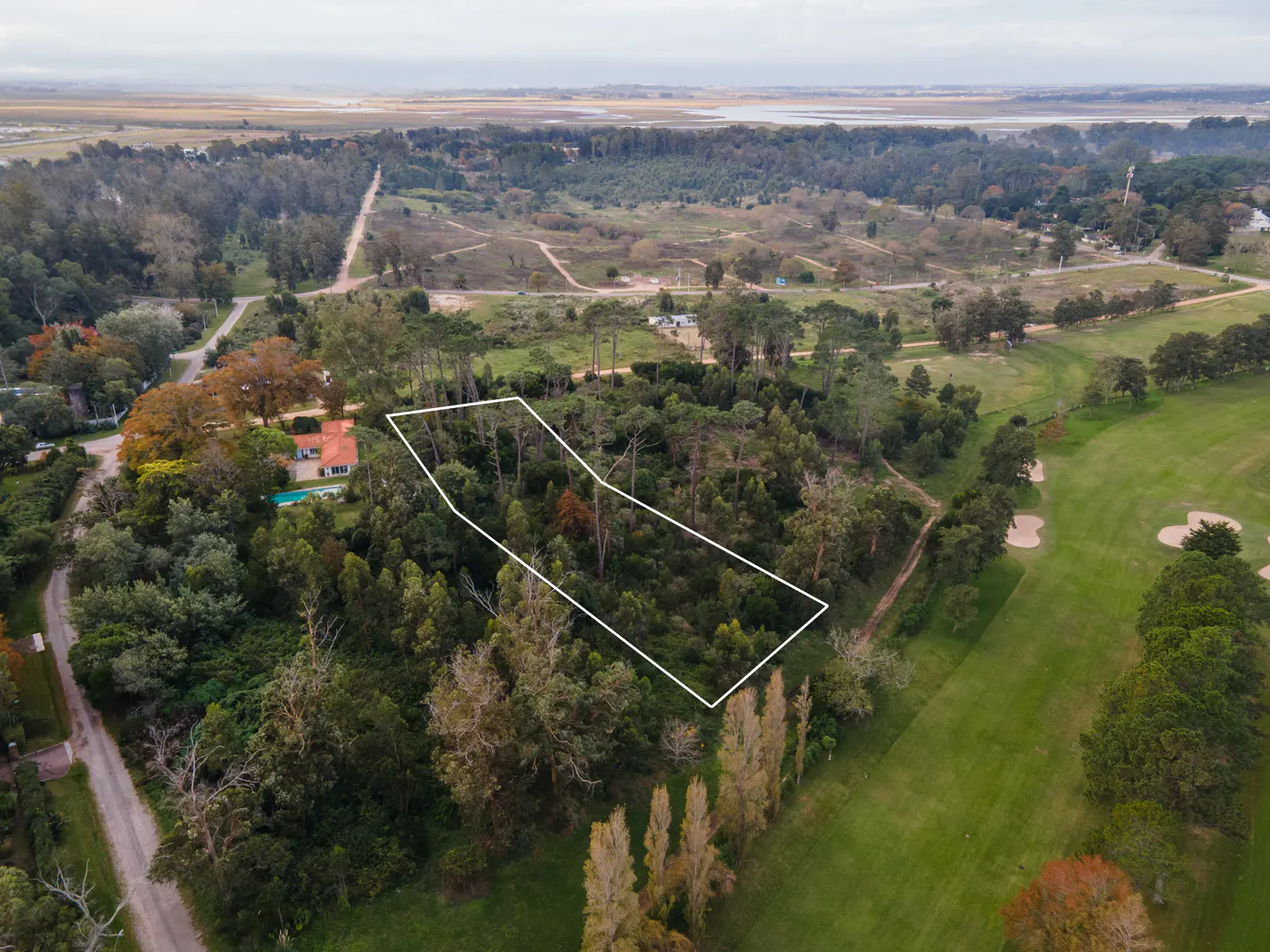 Aerial view of a wooded lot outlined in white, adjacent to a golf course and a house with a pool.