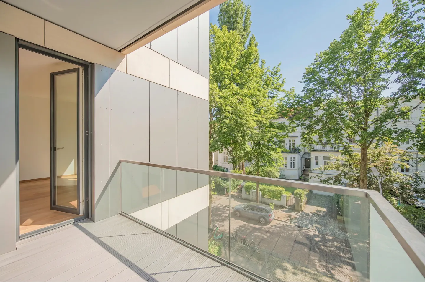 Balcony view with glass railing, gray wood floor, and open door to a bright room. Trees and a building are visible outside.