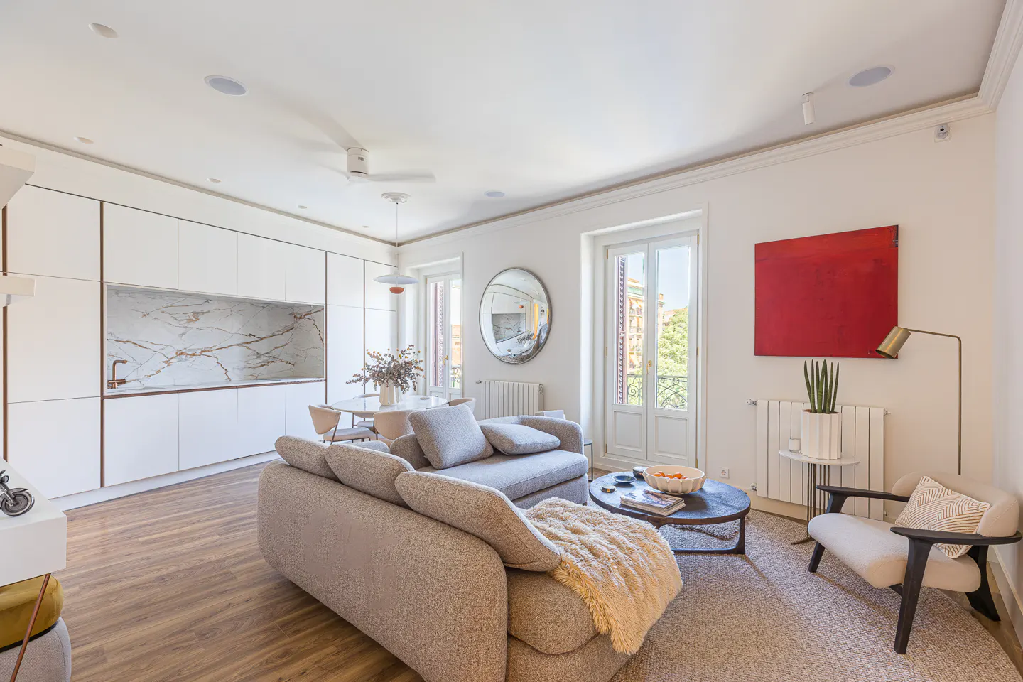 Bright, modern living room with a gray sofa, round table, and white kitchen cabinets with marble backsplash. A red painting hangs on the wall.