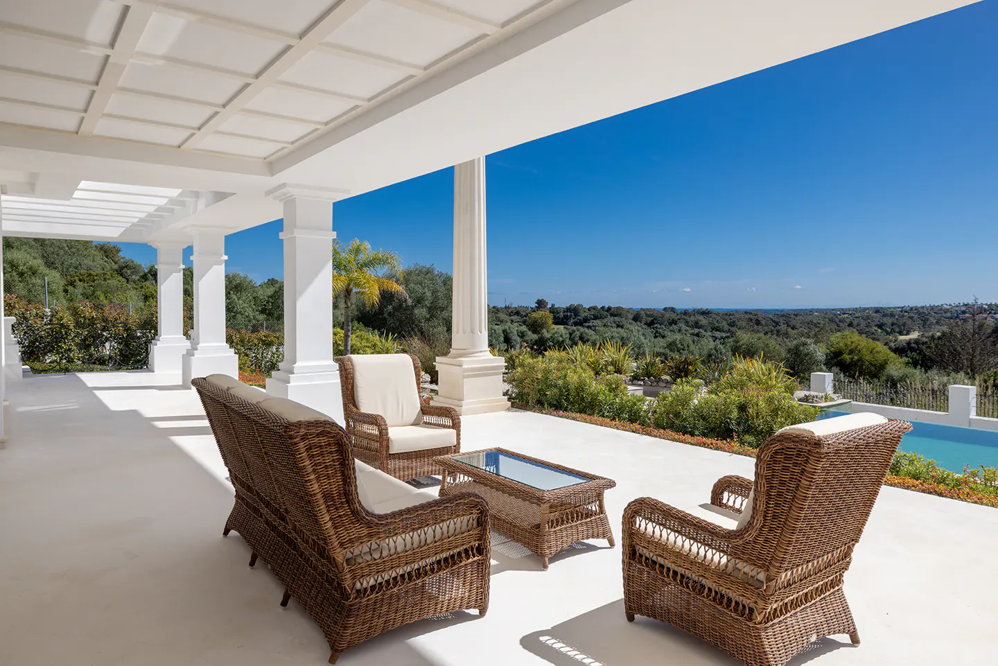 Outdoor patio with wicker furniture, white columns, and a view of lush greenery and a blue sky.