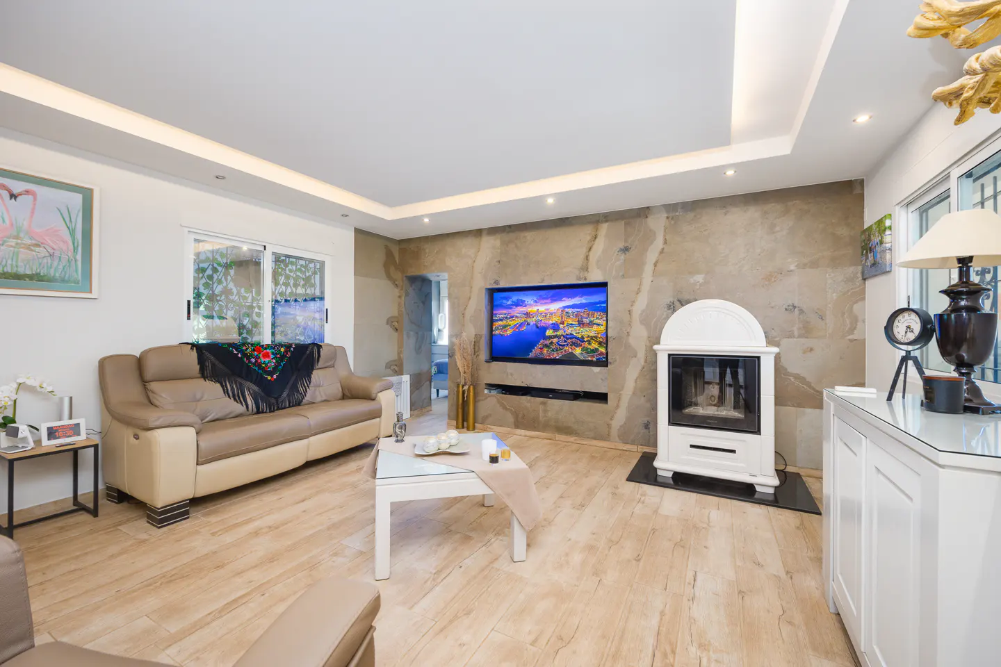 Living room with a tan leather sofa, white fireplace, and a flat-screen TV mounted on a stone wall. A white coffee table sits in the center.
