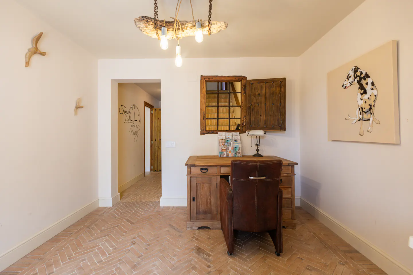 A rustic office space with a wooden desk, brown leather chair, and herringbone brick floor. A Dalmatian painting hangs on the wall.