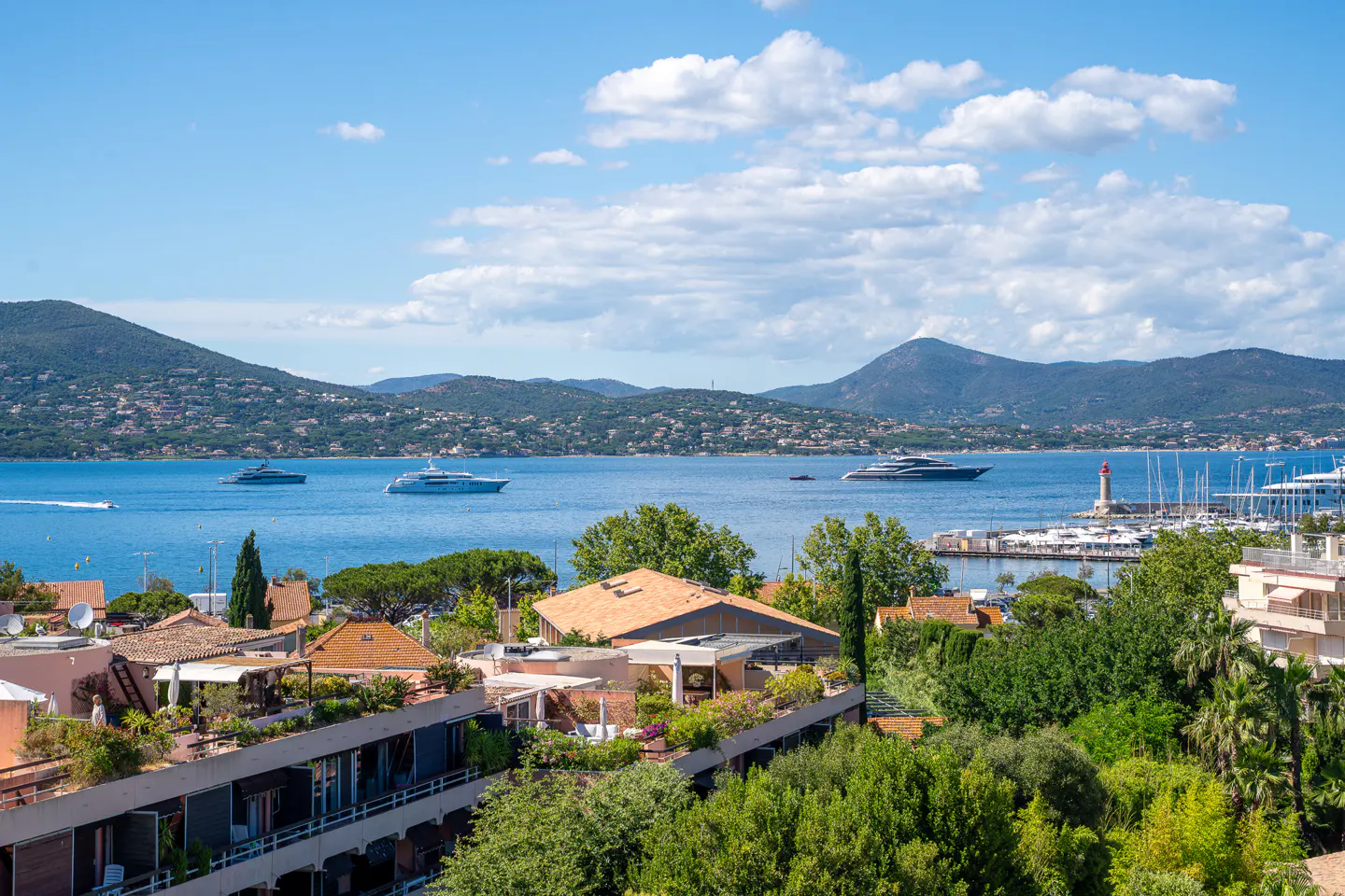 Elevated view of a coastal town with terracotta roofs, lush trees, and yachts on a blue sea under a sunny, cloudy sky.