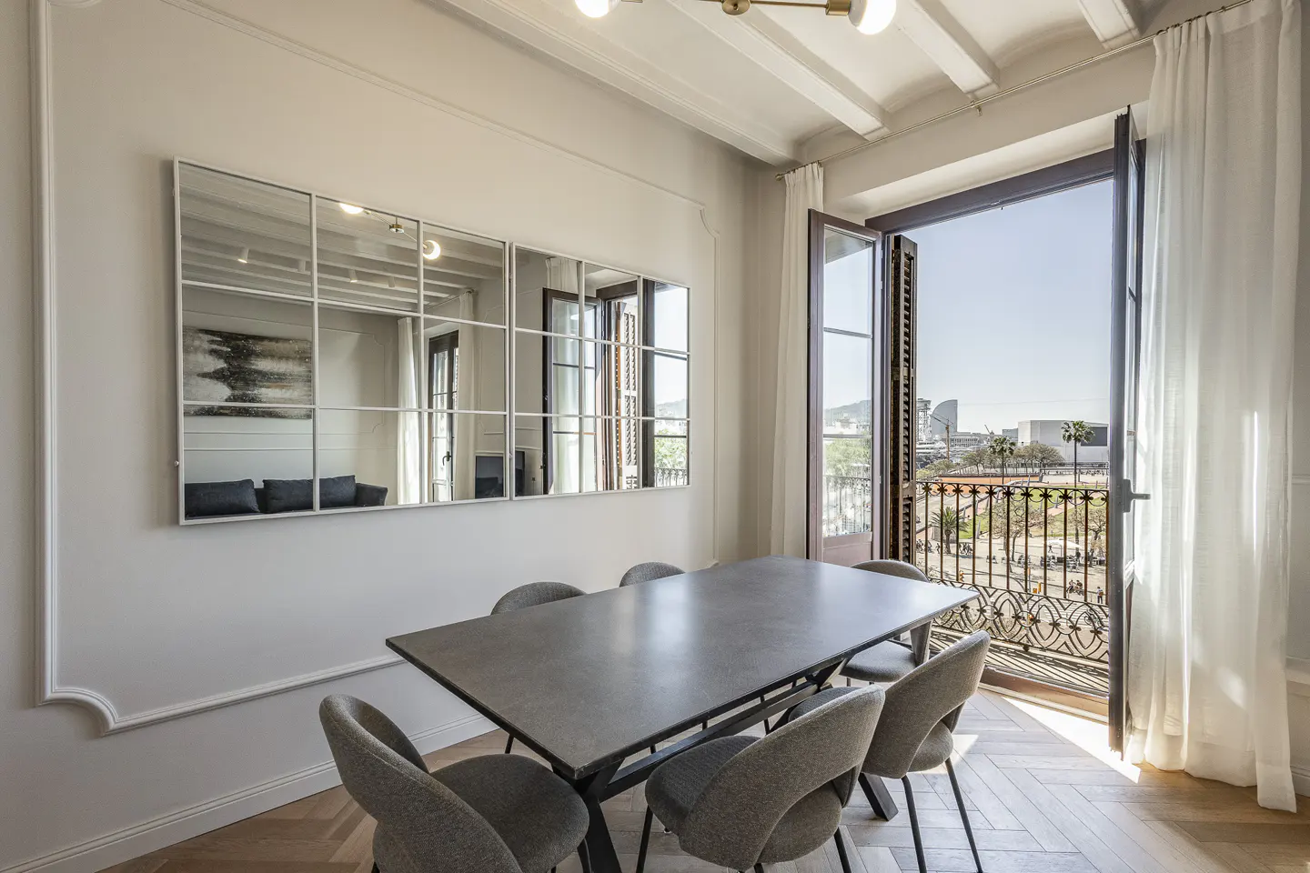 Bright dining room with a gray table, six chairs, and a large mirror. Open balcony doors reveal a city view.