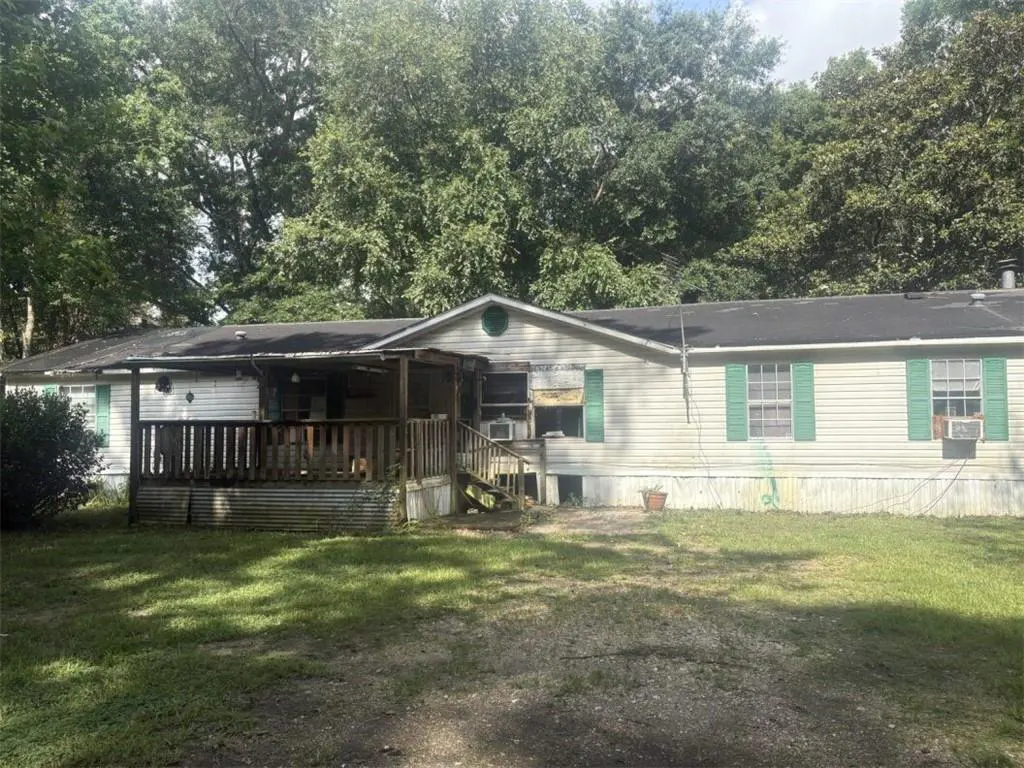 Single-story mobile home with white siding, green shutters, and a wooden front deck, set against a backdrop of tall trees.