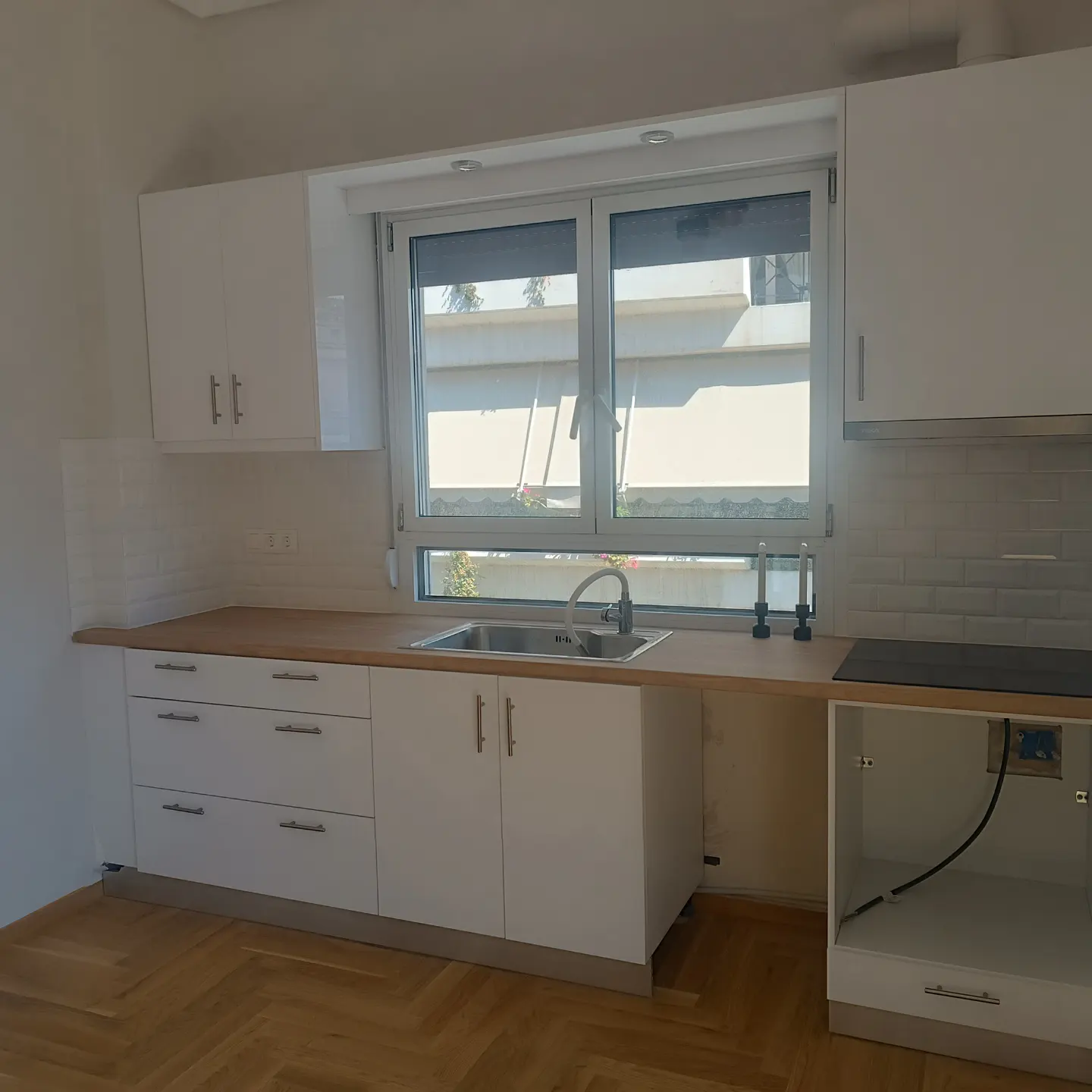 Bright kitchen with white cabinets, wood countertop, and herringbone wood floor. A window above the sink offers a view of the outside.