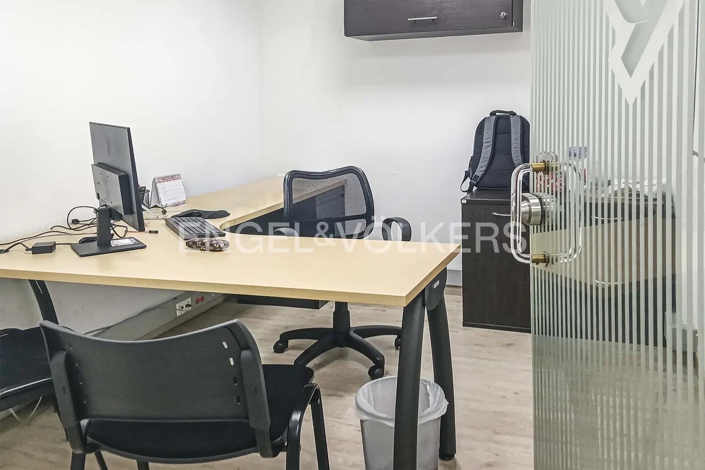 Office space with a light wood desk, black chairs, computer, and a gray backpack on a dark cabinet. A glass door is on the right.