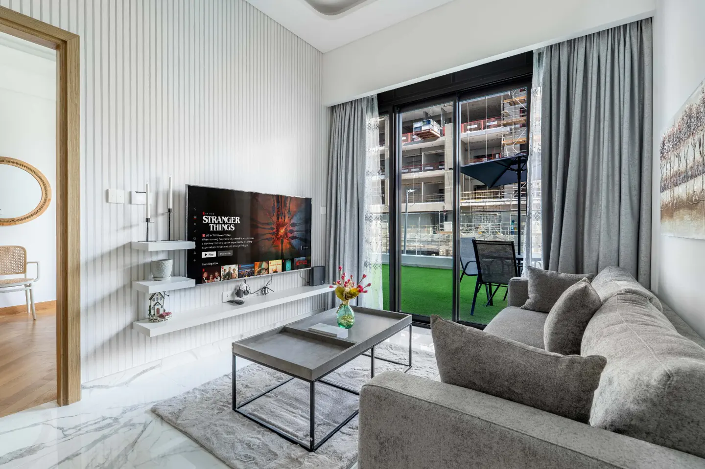 Modern living room with a grey sofa, coffee table, wall-mounted TV, and a balcony view of a building under construction.